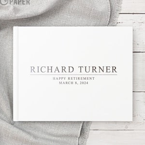 May include: White hardcover book with the text "RICHARD TURNER" in silver lettering. Below the name is the text "HAPPY RETIREMENT" and "MARCH 8, 2024" in smaller silver lettering.