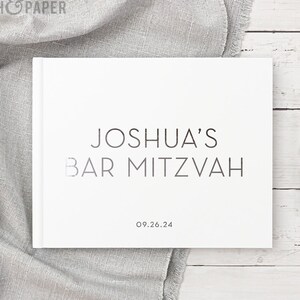 May include: White guest book with the text "Joshua's Bar Mitzvah 09.26.24" printed in silver foil.