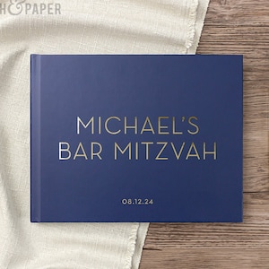 May include: A navy blue book with gold lettering that reads "MICHAEL'S BAR MITZVAH" and the date "08.12.24". A gold pen rests on a wooden surface next to the book. The book is on a cream-colored cloth.
