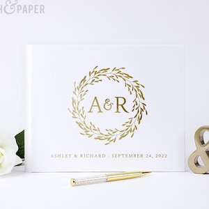 May include: White wedding guest book with a gold foil wreath design and the initials "A&R" in the center. The text "Ashley & Richard - September 24, 2022" is printed below the wreath.