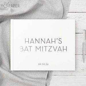 May include: A white hardcover book with the text "HANNAH'S BAT MITZVAH" in a minimalist font. The date "09.30.24" is below the text. A silver pen with a crystal-like design rests on a white wooden surface.