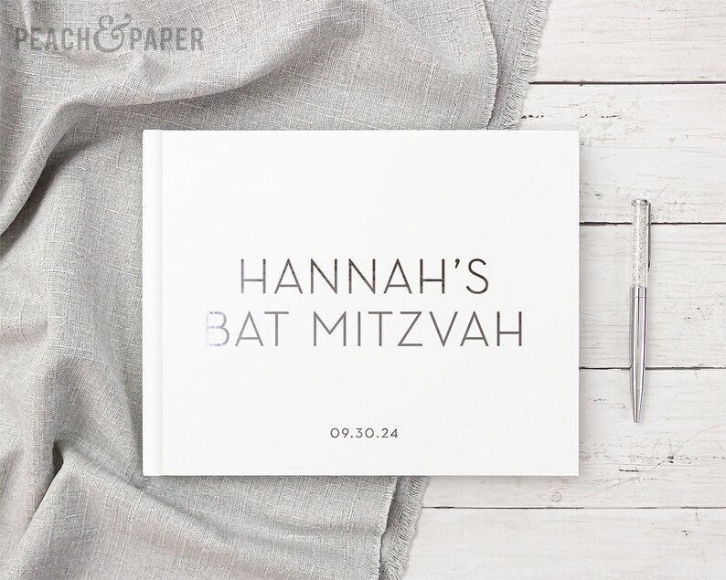 May include: A white hardcover book with the text "HANNAH'S BAT MITZVAH" in a minimalist font. The date "09.30.24" is below the text. A silver pen with a crystal-like design rests on a white wooden surface.