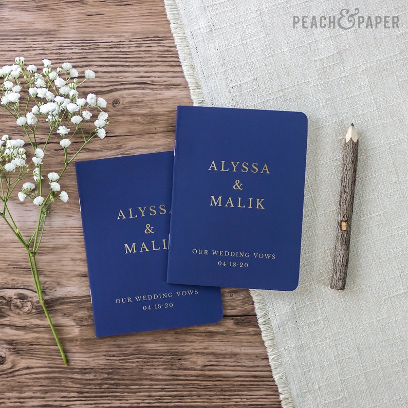 May include: Two navy blue wedding vow books with gold lettering. The books have the names Alyssa & Malik and the date 04-18-20 printed on the front. The books are on a wooden surface with a white fabric and a wooden pencil.