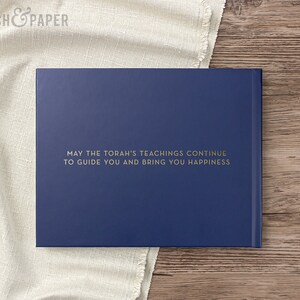 May include: A navy blue book with gold lettering that reads "MAY THE TORAH'S TEACHINGS CONTINUE TO GUIDE YOU AND BRING YOU HAPPINESS." The book is on a wooden surface with a white cloth.