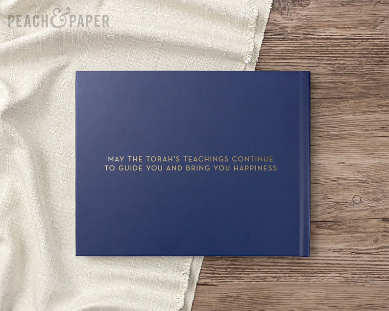 May include: A navy blue book with gold lettering that reads "MAY THE TORAH'S TEACHINGS CONTINUE TO GUIDE YOU AND BRING YOU HAPPINESS." The book is on a wooden surface with a white cloth.
