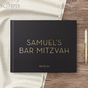 May include: A black guest book with gold lettering that reads "SAMUEL'S BAR MITZVAH" and the date "08.29.24". A gold pen with a glittery silver top rests on a wooden surface next to the book. A white cloth is in the background.