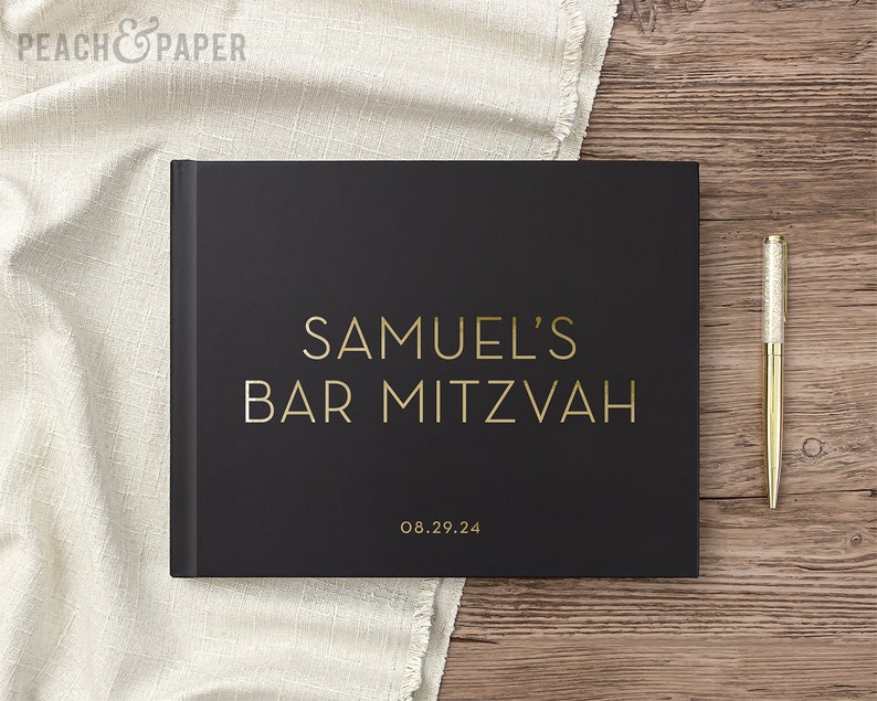 May include: A black guest book with gold lettering that reads "SAMUEL'S BAR MITZVAH" and the date "08.29.24". A gold pen with a glittery silver top rests on a wooden surface next to the book. A white cloth is in the background.