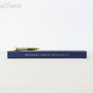 May include: A gold and silver pen rests on a navy blue box with the name "MICHAEL DAVID BENOWITZ" in gold lettering. The pen has a gold tip and a silver, glitter-filled barrel. The box is rectangular and has a simple design.