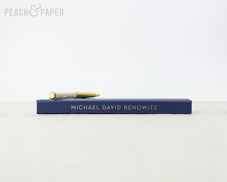 May include: A gold and silver pen rests on a navy blue box with the name "MICHAEL DAVID BENOWITZ" in gold lettering. The pen has a gold tip and a silver, glitter-filled barrel. The box is rectangular and has a simple design.
