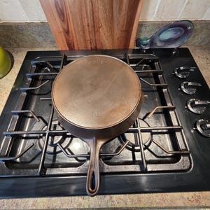 May include: A well-worn, cast iron skillet with a long handle, resting on a black gas stovetop. The skillet is a warm brown color, with the number "10" visible. The stovetop has silver burner grates and control knobs.