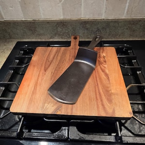 May include: A wooden chopping board with a handle and a stainless steel spatula on a black hob. The square wooden board is reddish-brown. The spatula is angled across the board. The hob has four burners.