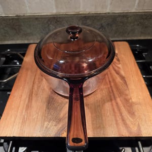 May include: A clear glass saucepan with a matching lid and a long handle, resting on a wooden cutting board. The glass has a brown tint. The saucepan is sitting on a black stovetop.