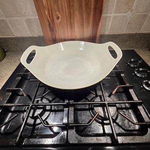 Op de afbeelding: Een crèmekleurige, ovaalvormige kookschotel met twee handvatten, rustend op een zwart gasfornuis. Een houten snijplank staat op de achtergrond. Een groene kom is links zichtbaar.