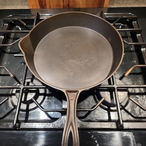 May include: A dark, cast iron skillet with a long handle and a rounded cooking surface. The skillet is sitting on a black stovetop with a gas burner. The handle has a hole at the end for hanging. The skillet is approximately 10 inches in diameter.