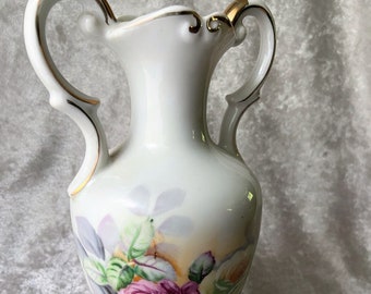 May include: White porcelain vase with two handles and gold trim. The vase is decorated with a floral pattern of pink, white, and yellow roses.