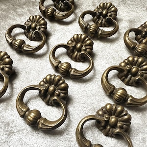 May include: A collection of ten brass-toned metal drawer pulls, each featuring a circular backplate with a floral-inspired petal design and a swinging, curved handle with a central ribbed detail, arranged in a grid pattern on a surface.