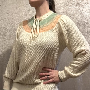 May include: Cream-colored knit sweater with a textured pattern. The neckline features a color block design in shades of green and orange, with a cream-colored tie. The long sleeves have ribbed cuffs. A vintage-inspired top.