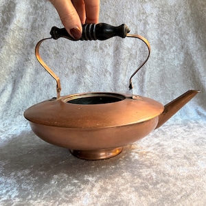 Puede incluir: Una tetera de cobre con un asa de madera. La tetera tiene un cuerpo ancho y poco profundo y un pico en el lateral.