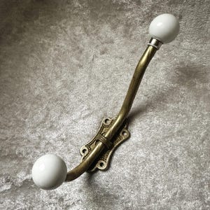 以下が含まれることがあります： アンティーク真鍮仕上げの金属製アームと装飾的な取り付けプレートを備えた壁掛けフック。フックの両端には滑らかで丸い白いセラミック製のノブが付いており、衣類やアクセサリーを掛けるための機能的なデザインになっています。