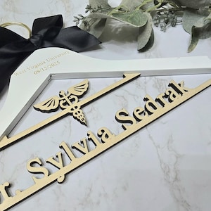 May include: White wooden hanger with a black satin bow and gold-tone metal hook. The hanger is engraved with "West Virginia University 09/12/2025". Two wooden nameplates read "Dr. Sylvia Sedrak" and feature a medical symbol.