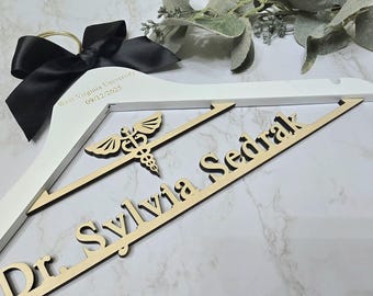 May include: A white wooden clothes hanger featuring a black satin bow at the top. The hanger is personalized with the text West Virginia University 09/12/2025, a wooden caduceus medical symbol, and the name Dr. Sylvia Sedrak in large wooden letters.