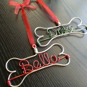 May include: Two silver bone-shaped ornaments with the names "Bella" and "Duke" written in red and green wire, respectively. The ornaments are hanging from red and green ribbons.