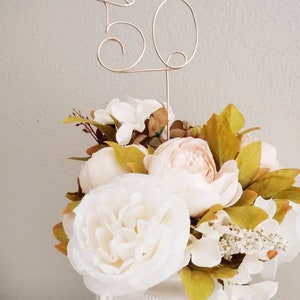 May include: A gold wire table number with the number "50" in a decorative font. The number is placed in a white vase with a bouquet of white and peach peonies and greenery.