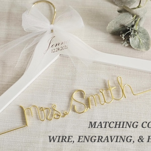 May include: A white wooden clothes hanger featuring a gold wire script reading Mrs. Smith, a white tulle bow tied to the gold hook, and the engraved name Jenna with the date 12.24.2025. Text on the image reads MATCHING COLOR WIRE, ENGRAVING, & HOOK.