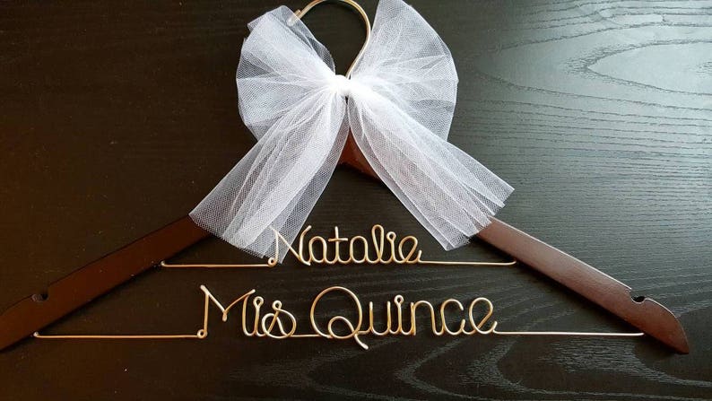 Pu&ograve; includere: Un appendiabiti in legno marrone con scritte dorate "Natalie" e "Mis Quince". Un fiocco di tulle bianco &egrave; attaccato in cima. L'appendiabiti &egrave; su una superficie scura.