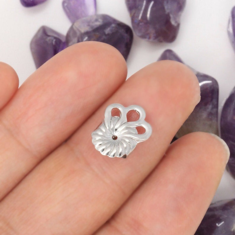 Peut inclure: Un fermoir de boucle d'oreille argent&eacute; pr&eacute;sentant un design en &eacute;ventail festonn&eacute; avec deux boucles ouvertes sur le dessus. Le composant m&eacute;tallique poss&egrave;de un trou central pour la tige et un motif rayonnant textur&eacute; sur sa surface.
