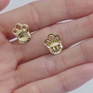 May include: Two gold-toned earring backs featuring a decorative, scalloped, crown-like top edge with a textured, fan-shaped base. These metal earring fasteners are designed to provide support and security for post-style earrings.