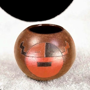 May include: A round, brown ceramic vessel with a dark interior. The exterior features a stylized face design in red, black, and brown. The vessel has a textured surface and a wide opening. The design is symmetrical and geometric.