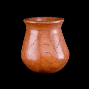 Puede incluir: Una pequeña olla de cerámica marrón con cuerpo redondeado y borde ligeramente acampanado. La olla está decorada con un diseño simple pintado a mano con líneas marrones.