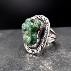 May include: A silver ring featuring a large, irregular, textured green turquoise stone set in a bezel. The silver band has a wide, flat face decorated with a repeating stamped cross pattern around the perimeter of the central stone setting.