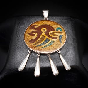 May include: A circular silver pendant featuring a central inlay of speckled tan stone with a dark brown and blue geometric design. Four smooth, teardrop-shaped silver dangles are suspended from the bottom edge of the circular frame by small rings.