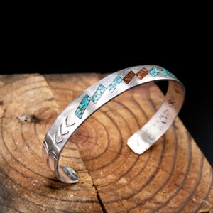 May include: A silver cuff bracelet featuring a zigzag inlay pattern of small turquoise and reddish-brown stone fragments. The metal surface is engraved with repeating chevron-like markings along the side, creating a textured appearance.