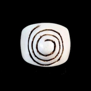 Puede incluir: Un anillo blanco con un diseño de espiral marrón oscuro. El anillo tiene forma rectangular con esquinas redondeadas. La espiral está centrada, creando un contraste llamativo sobre el fondo blanco.