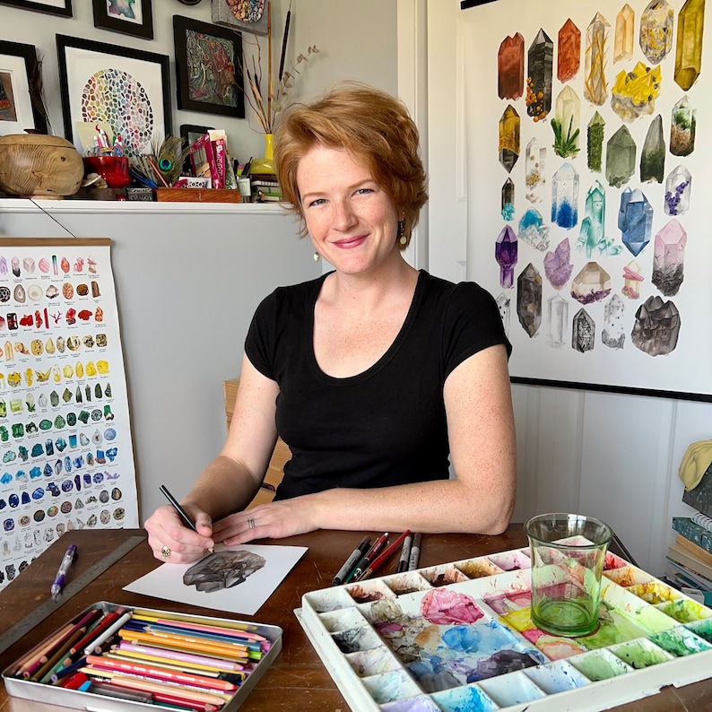 May include: A woman with red hair sits at a table with a watercolor palette, paintbrushes, and colored pencils. She is drawing a crystal on a piece of paper. There is a chart of crystals on the wall behind her.