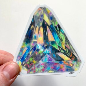 May include: A colorful, geometric, triangular sticker with a detailed drawing of a crystal. The crystal is drawn in shades of blue, green, yellow, and purple. The sticker has a white background and a black signature in the bottom right corner.