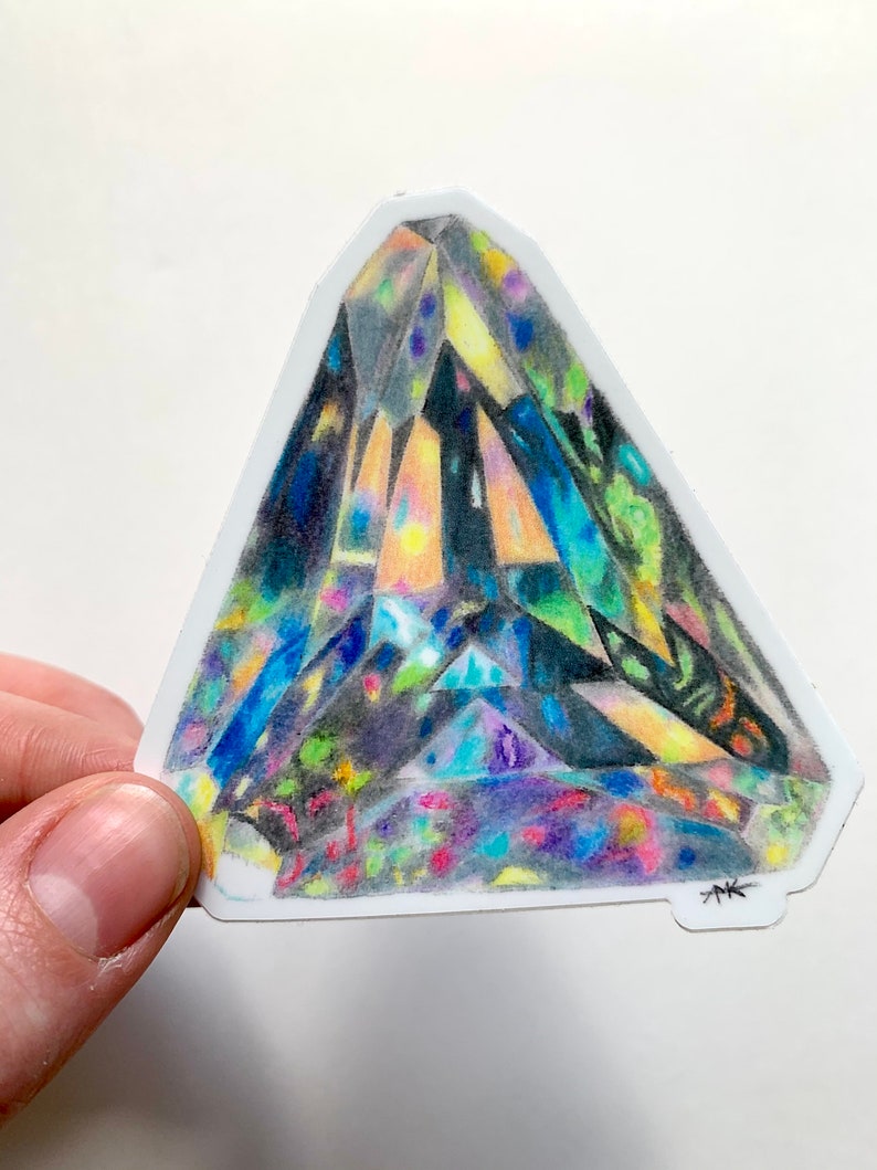 May include: A colorful, geometric, triangular sticker with a detailed drawing of a crystal. The crystal is drawn in shades of blue, green, yellow, and purple. The sticker has a white background and a black signature in the bottom right corner.