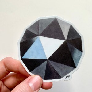 May include: A black and white geometric sticker of a diamond with a blue center. The sticker has a white border and the artist's initials "AK" are in the bottom right corner.