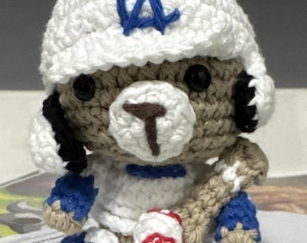May include: Crocheted bear wearing a white baseball cap with a blue "LA" logo. The bear is dressed in a white baseball uniform with blue accents and holds a baseball bat and ball. The bear is approximately 5 inches tall.