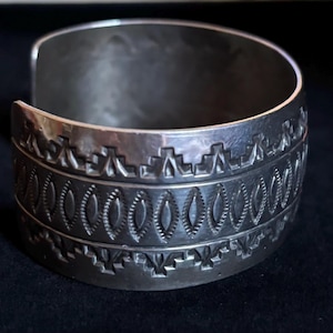 May include: A wide silver cuff bracelet featuring a polished metallic finish and a repeating stamped pattern. The design consists of a central row of vertical oval shapes flanked by two rows of geometric stepped motifs along the top and bottom edges.