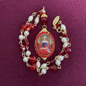 Può includere: Un pendente dorato con un'immagine religiosa, circondato da gemme rosse, è appeso a un braccialetto di perline. Il braccialetto presenta perline sfaccettate rosse, perle finte e maglie di catena dorate. Lo sfondo è bordeaux scuro.
