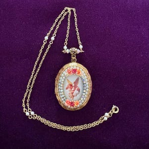 May include: A gold-toned oval locket necklace featuring a central image of a winged cherub. The locket is framed by small iridescent crystals and miniature pink and orange floral accents. The chain includes small white beads along its length.