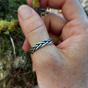 May include: A silver ring with a woven, braided design. The ring is worn on a finger, highlighting its detailed pattern. The metallic sheen of the ring contrasts with the natural background.