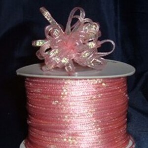 May include: A spool of pink ribbon with a decorative bow on top. The ribbon is a light pink color with a slight shimmer. The bow is made of the same ribbon material and is also pink. The spool is white.