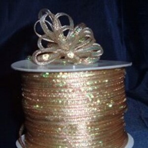 May include: A spool of metallic gold cord featuring a shimmering, iridescent finish. The cord is tightly wound around a white plastic cylinder, with a decorative bow made from the same cord resting on the top edge of the spool against a dark background.