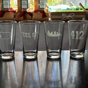 May include: Four clear pint glasses with etched designs. One glass features a heart within a tag shape, another reads "STEEL CITY", a third displays a city skyline, and the last has the number "412". The glasses are on a reflective surface.