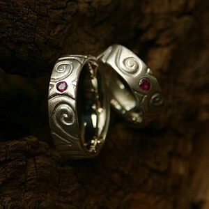 Könnte beinhalten: Zwei silberne Eheringe mit einem wirbelnden Design und einem kleinen roten Edelstein, der in jeden Ring eingefasst ist.
